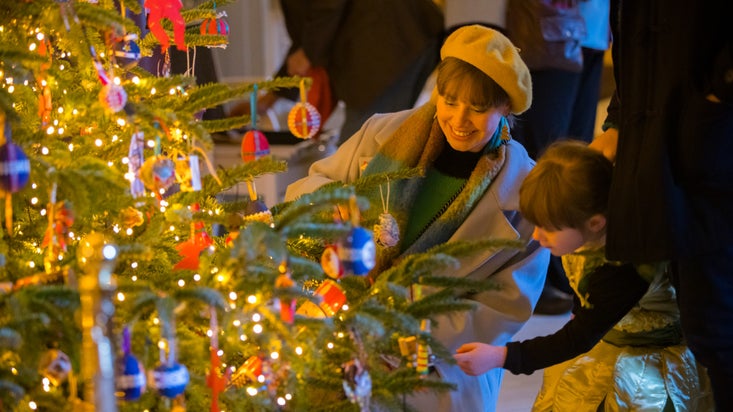 A woman in a pale grey winter coat and beret crouching down to look at a Christmas tree with a young girl in a black long sleeved top and gold dres